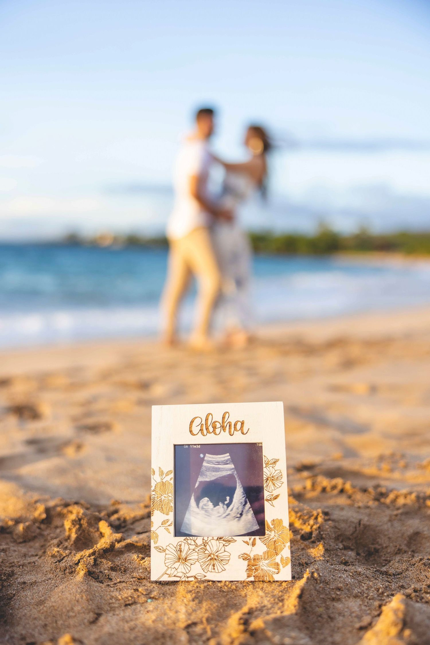 Framed ultrasound photo with Aloha on a sandy Hawaii beach, with an expecting couple embracing in soft focus by the ocean. Makena