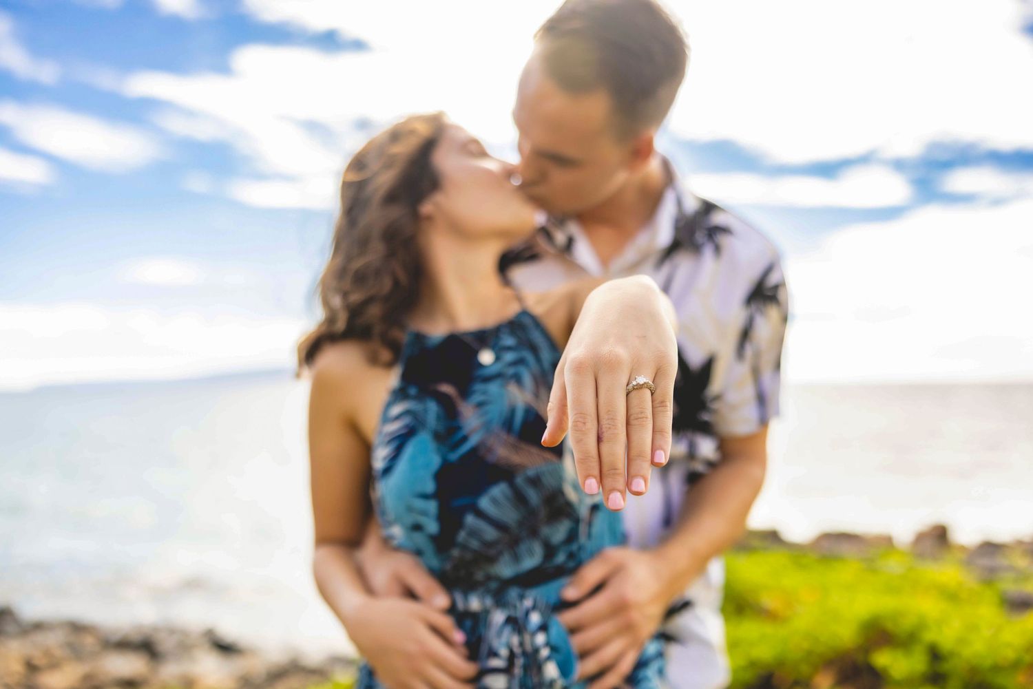 Couple kissing by the ocean as the woman shows off her new engagement ring in a tropical seaside proposal photo. Wailea
