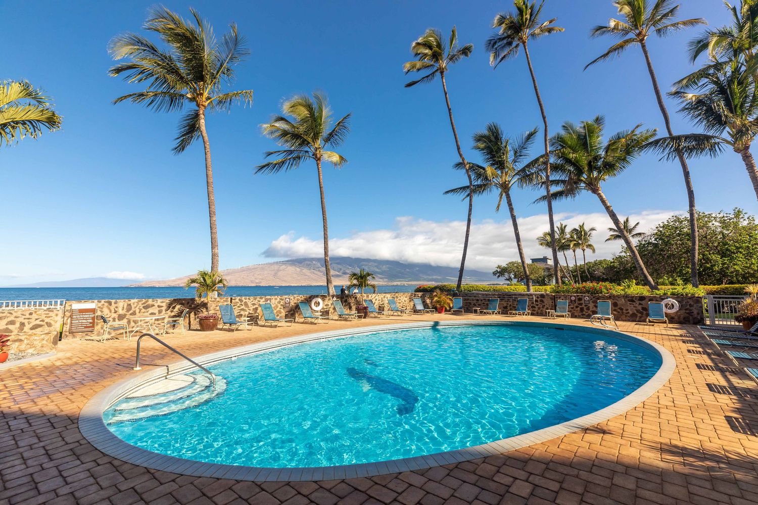 Tropical resort swimming pool with a brick paved deck, tall palm trees, lounge chairs, and ocean and mountain views under a blue sky.