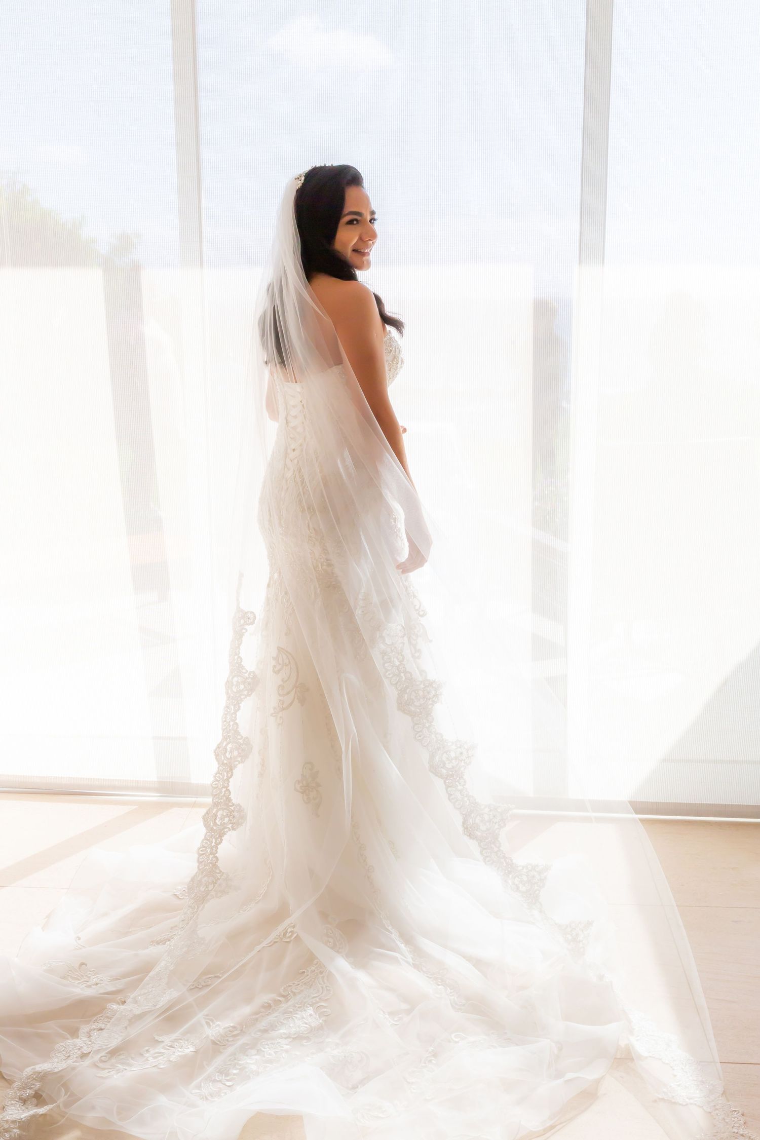 Bride in an elegant lace wedding gown with a long flowing veil and train smiles over her shoulder beside a bright sunlit window. Kahana
