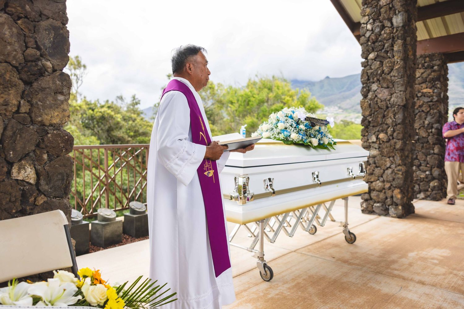 A priest in white vestments and a purple stole conducts an outdoor funeral service beside a white casket topped with blue flowers.