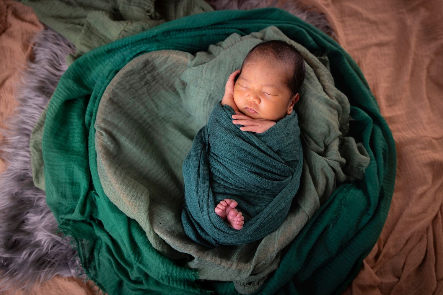 Sleeping newborn swaddled in a dark green wrap lies on layered green blankets and a fur backdrop.