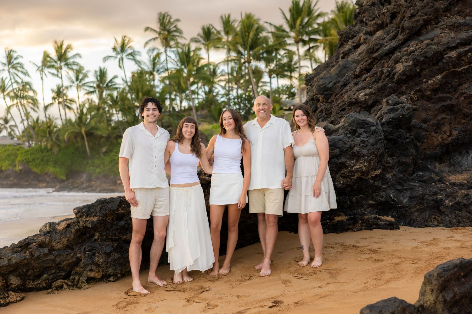 Family of five in white outfits stands barefoot on a sandy Hawaii beach beside lava rocks with palm trees behind them at sunset. Makena