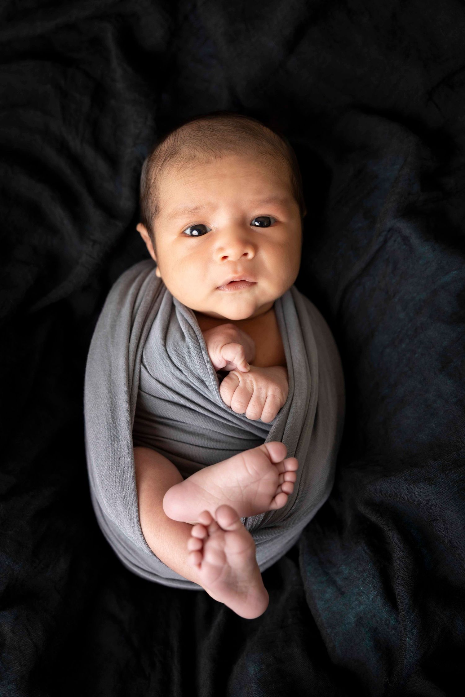 Alert newborn wrapped in a gray swaddle with tiny bare feet showing, lying on a dark black blanket.