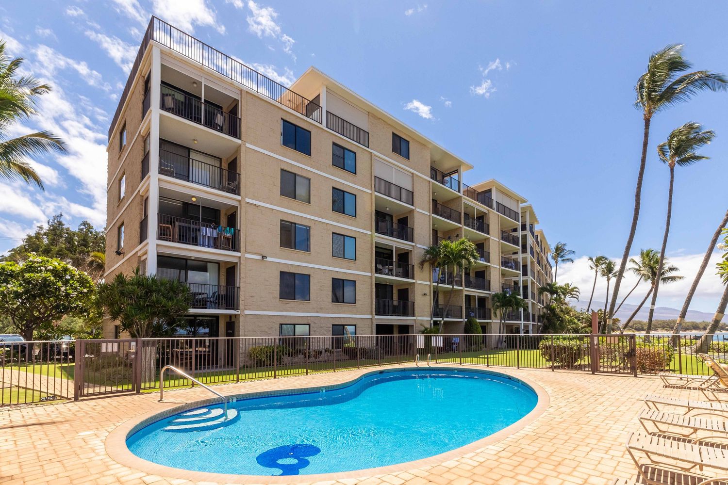 Multi-story oceanfront condo building with balconies beside a curved outdoor pool, palm trees, and lounge chairs under a bright blue sky.