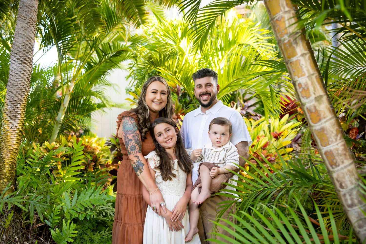 Smiling family of four with a young girl and baby posing together among lush tropical palms and colorful foliage in a outdoor portrait.
