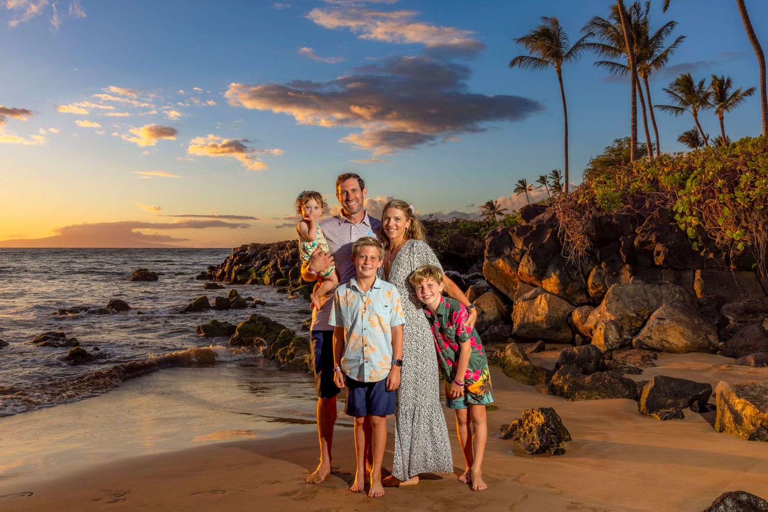 Family of five in coordinated outfits poses on a rocky tropical beach at sunset with palm trees and a colorful sky.