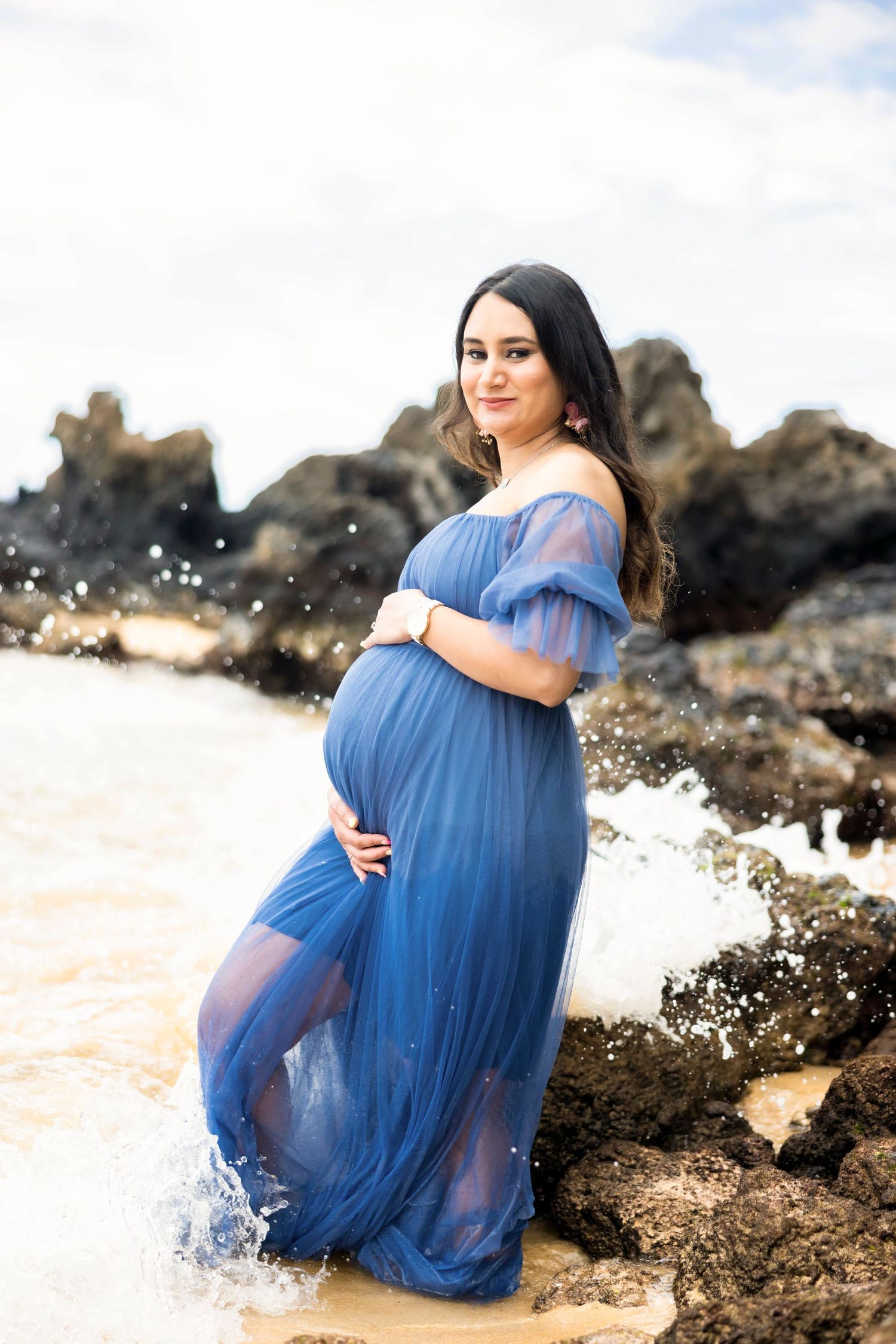 Pregnant woman in a flowing blue tulle maternity gown cradling her belly on a rocky beach as ocean waves splash behind her. Makena
