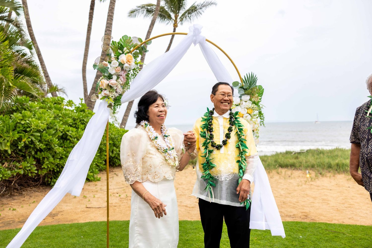Couple wearing green and floral leis stands beneath a white draped wedding arch with flowers on a grassy oceanfront in Hawaii. Kihei