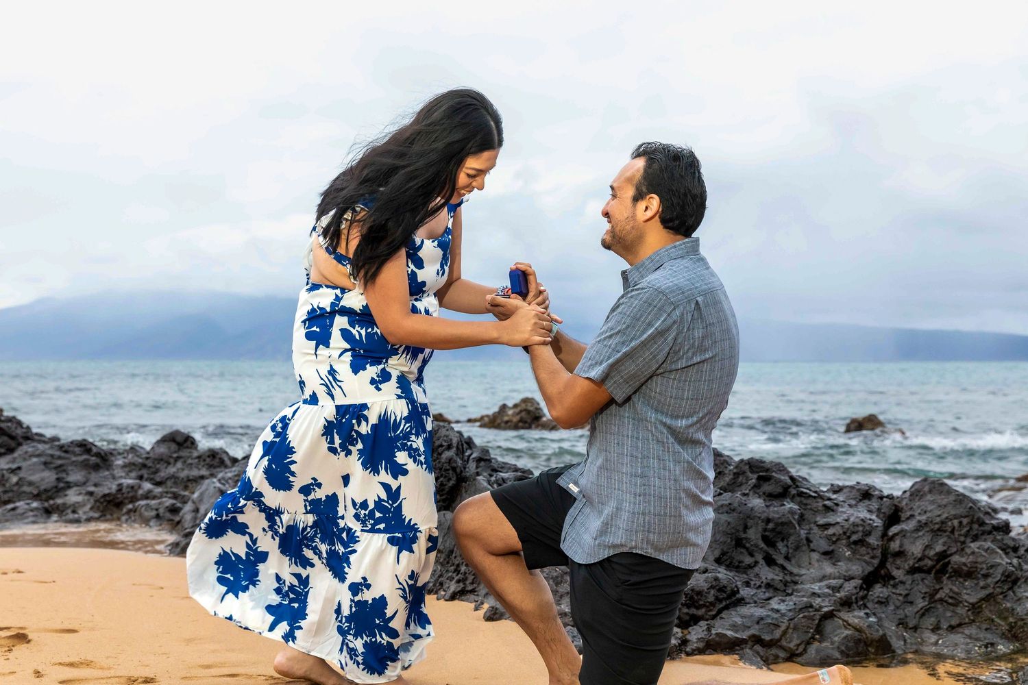 Man kneeling on a rocky beach proposes to a smiling woman in a blue floral dress, holding out a ring box by the ocean. Napili Bay