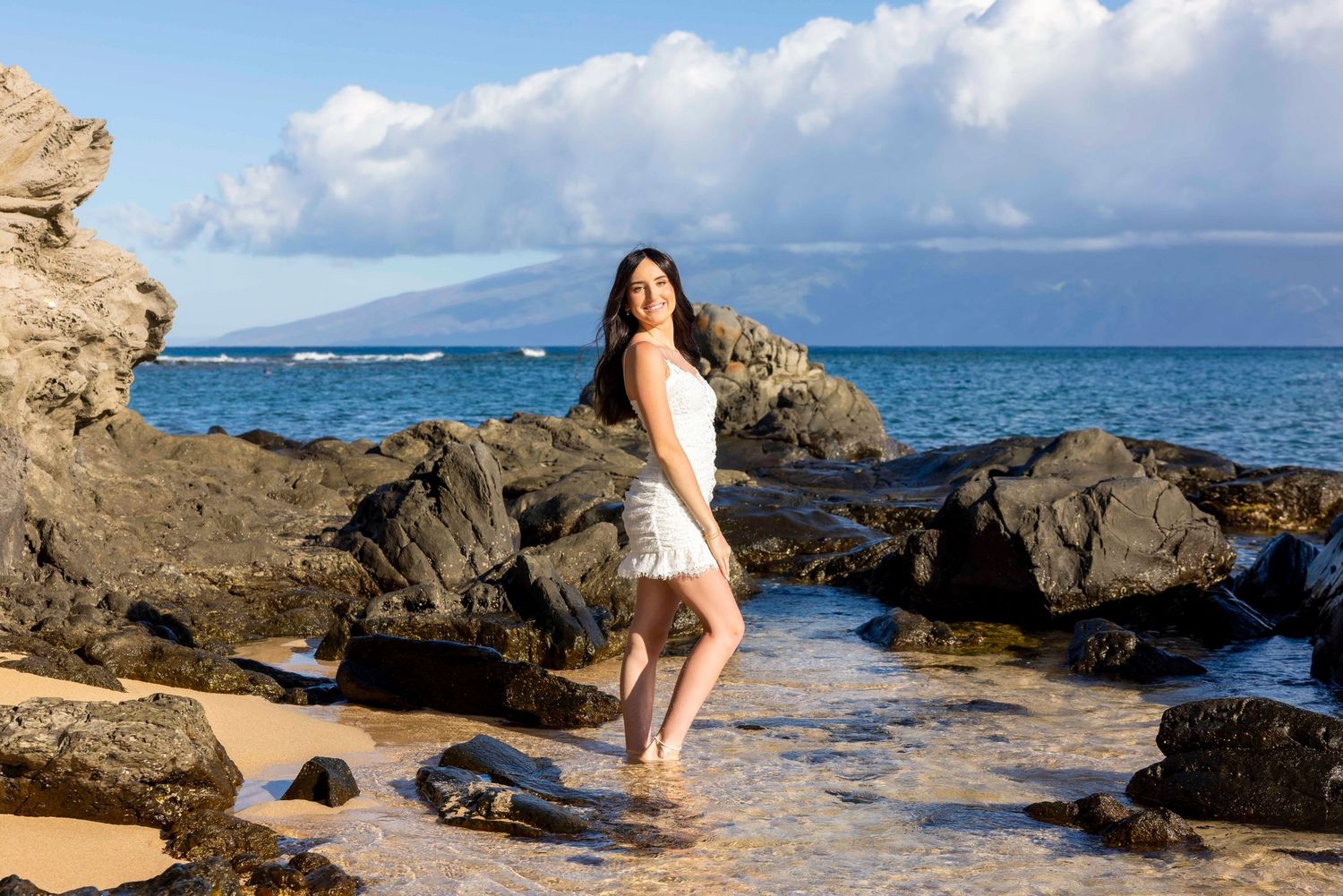 Woman in a white dress stands barefoot in shallow water among dark volcanic rocks on a tropical beach with ocean and distant mountains.