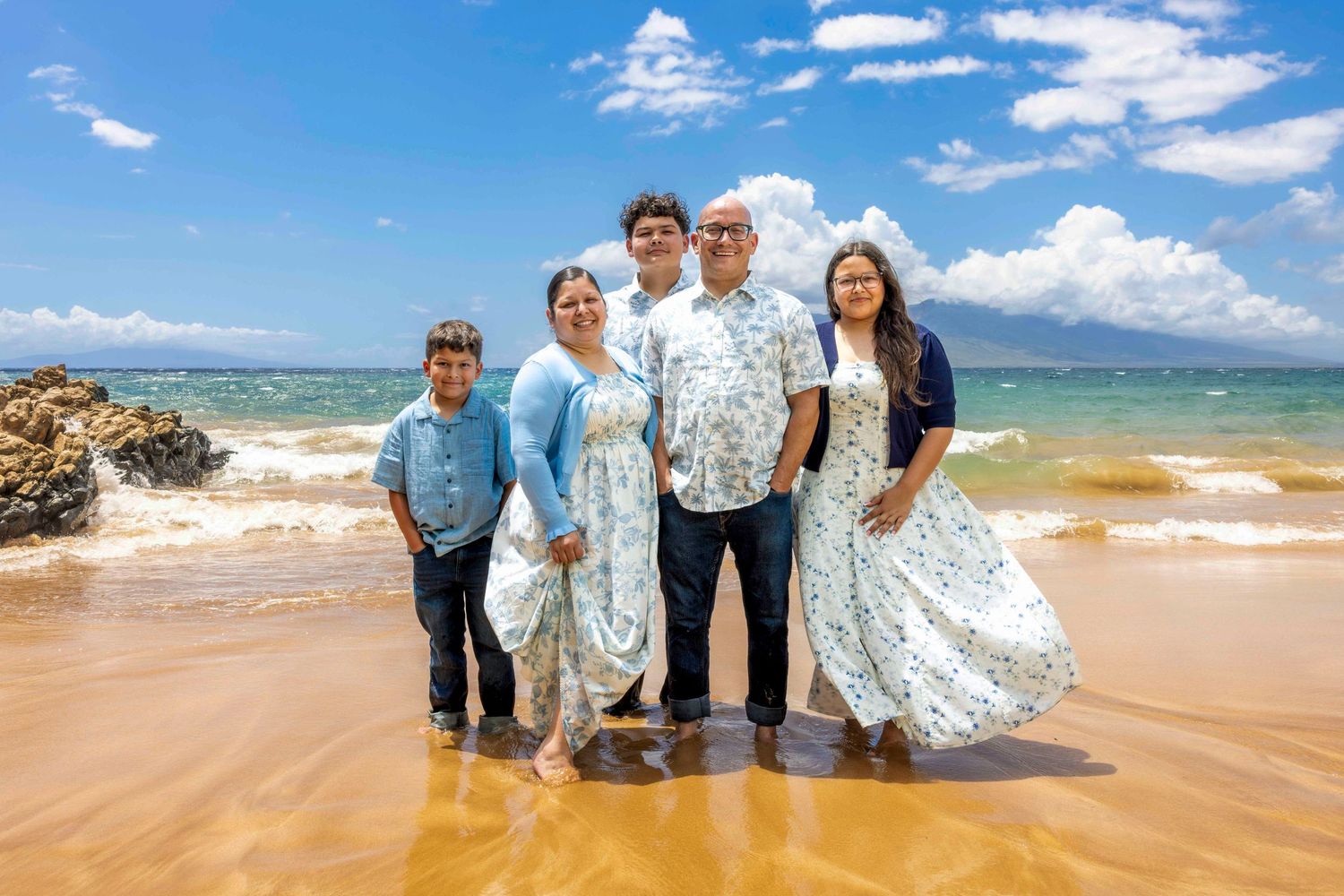 Family of five in matching floral outfits posing together on a wet sandy beach with ocean waves, rocks, and a blue sky with clouds.