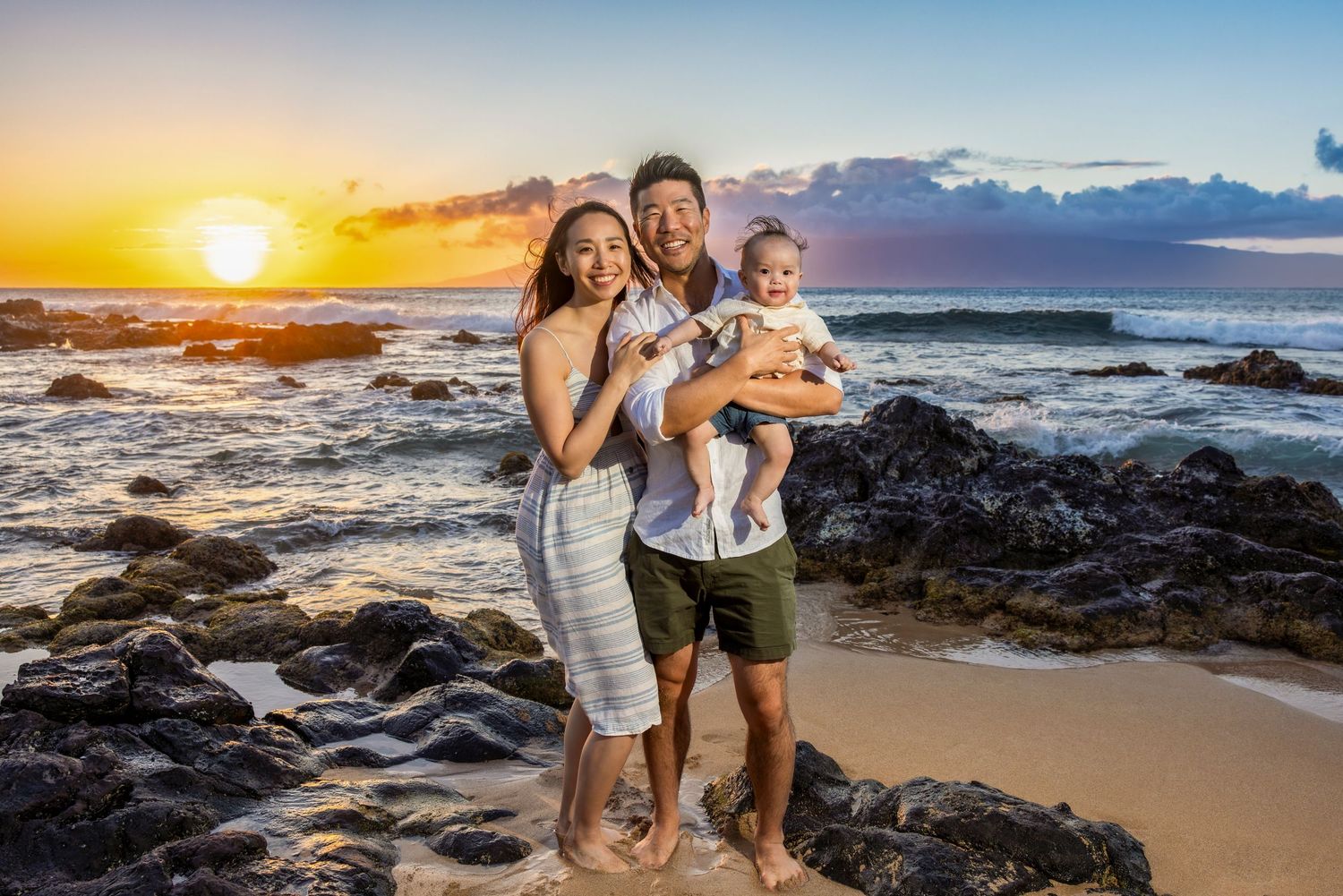 Couple holds their baby on a rocky Hawaii shoreline as the sun sets over the ocean in a warm family beach portrait.