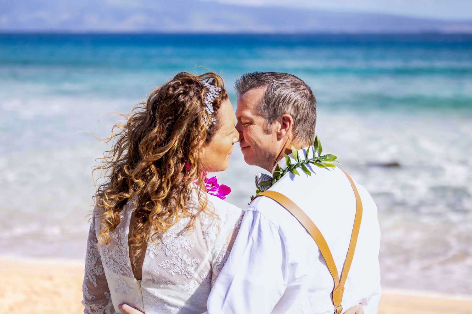 Bride and groom share a tender moment on a sunny Hawaiian beach, wearing a floral lei and suspenders with turquoise ocean waves behind them.