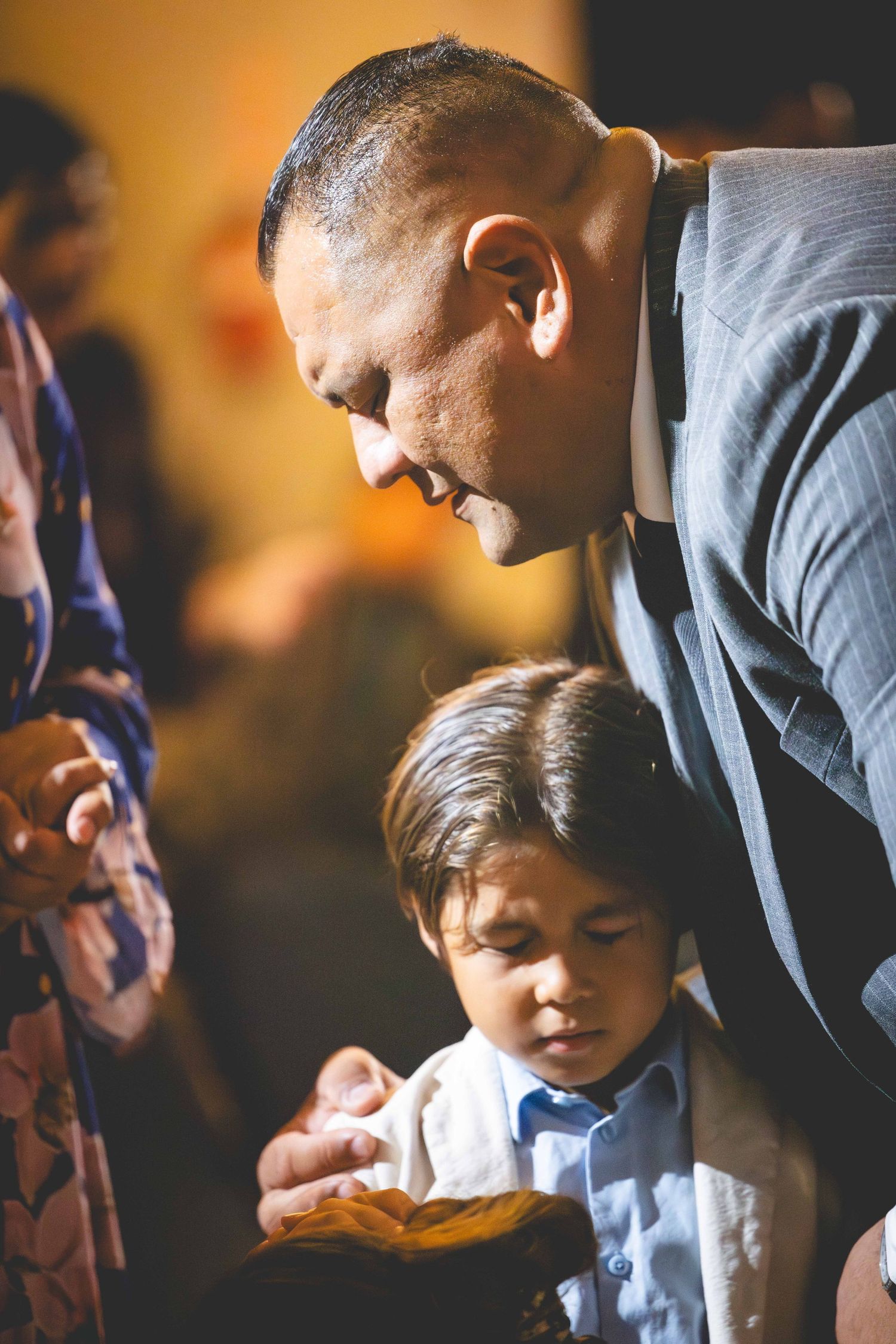 A man in a gray pinstripe suit bows his head and places a hand on a young boy's shoulder in a warm, candlelit moment.