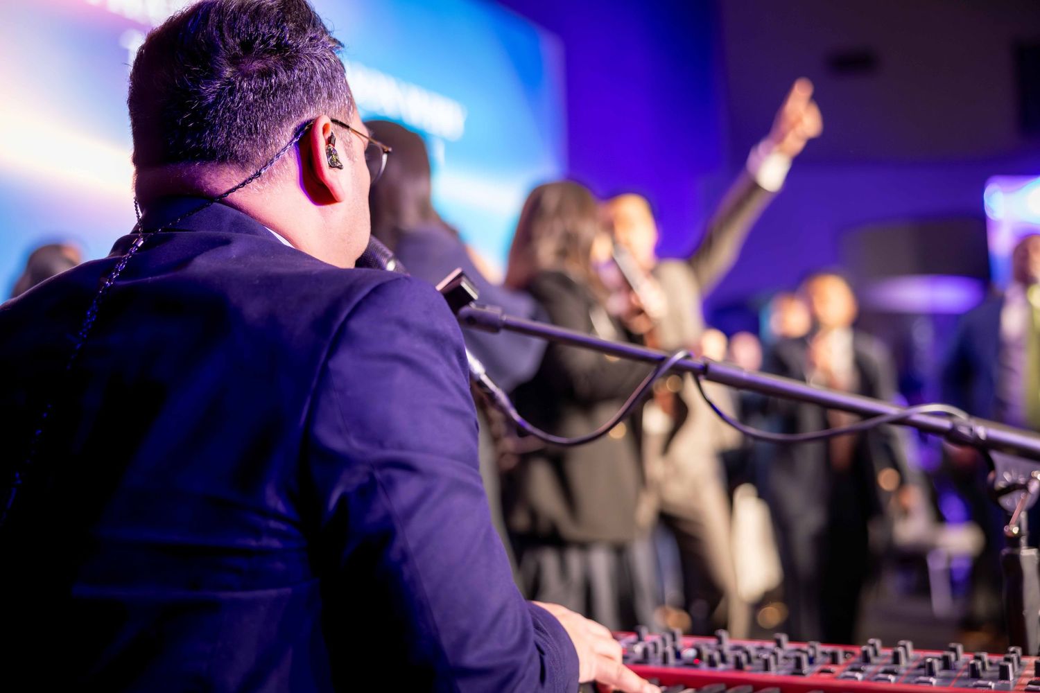 Musician in a dark suit plays a keyboard in front of a microphone while an enthusiastic crowd raises their hands at a live event.