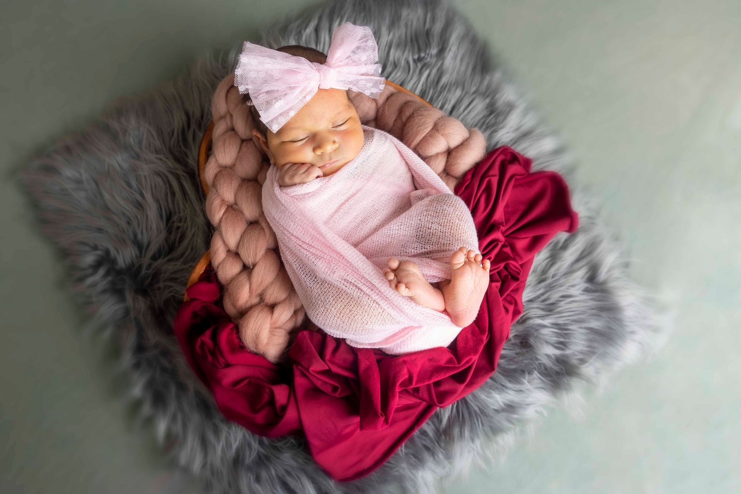 Newborn girl sleeping in a basket with a pink bow headband and pink wrap, surrounded by gray fur and a burgundy fabric.