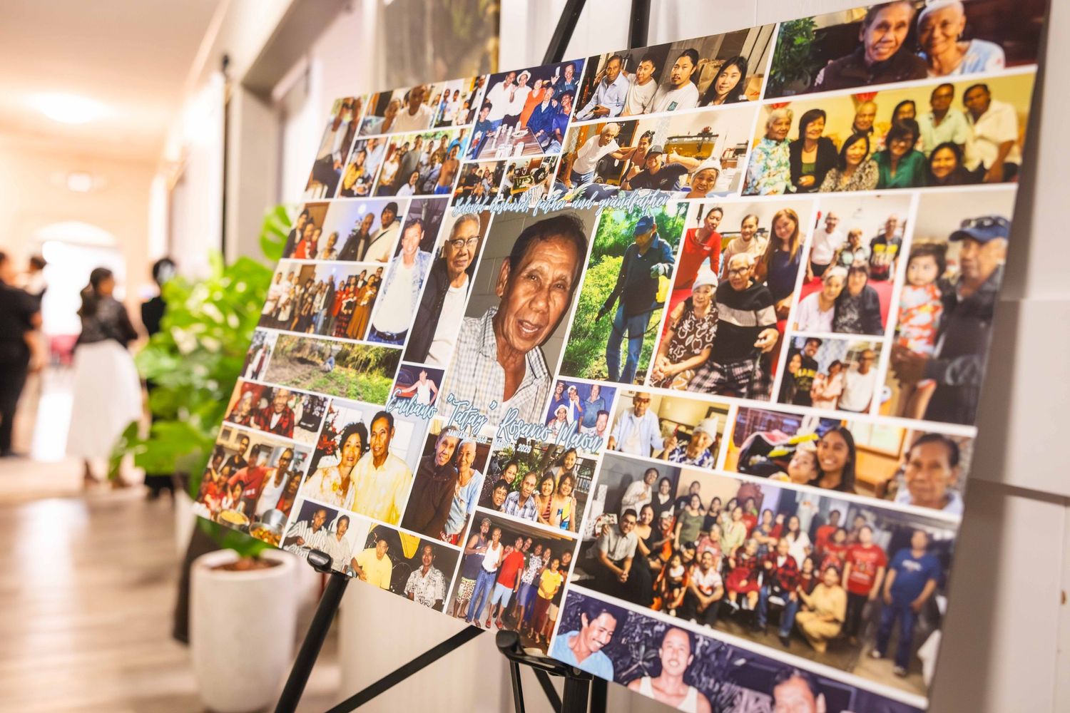 Large memorial photo collage of many smiling people displayed on an easel in a bright hallway with a potted plant nearby.