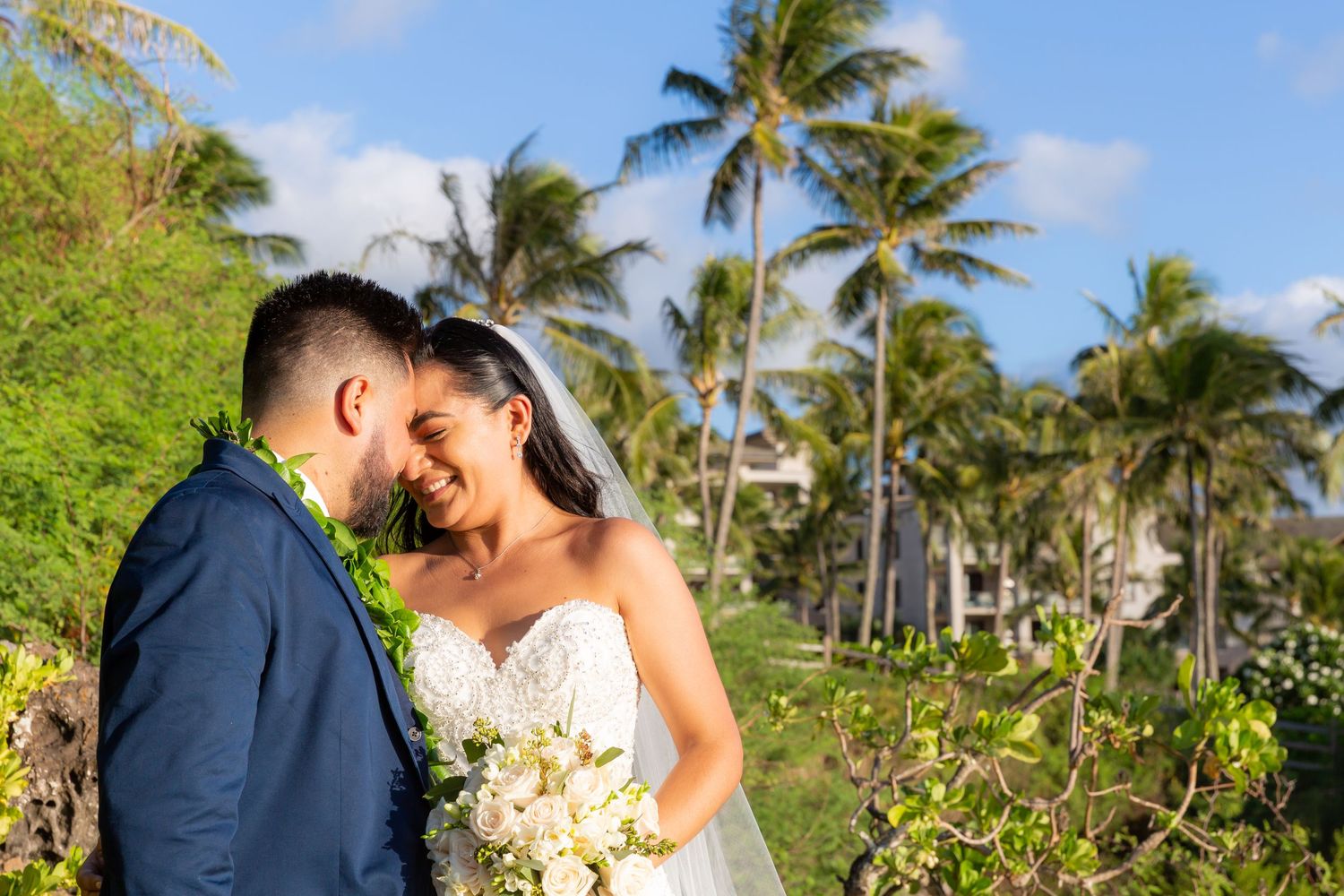 Smiling bride in a white lace gown and veil laughs with her groom in a navy suit and green lei among palm trees at a tropical wedding.