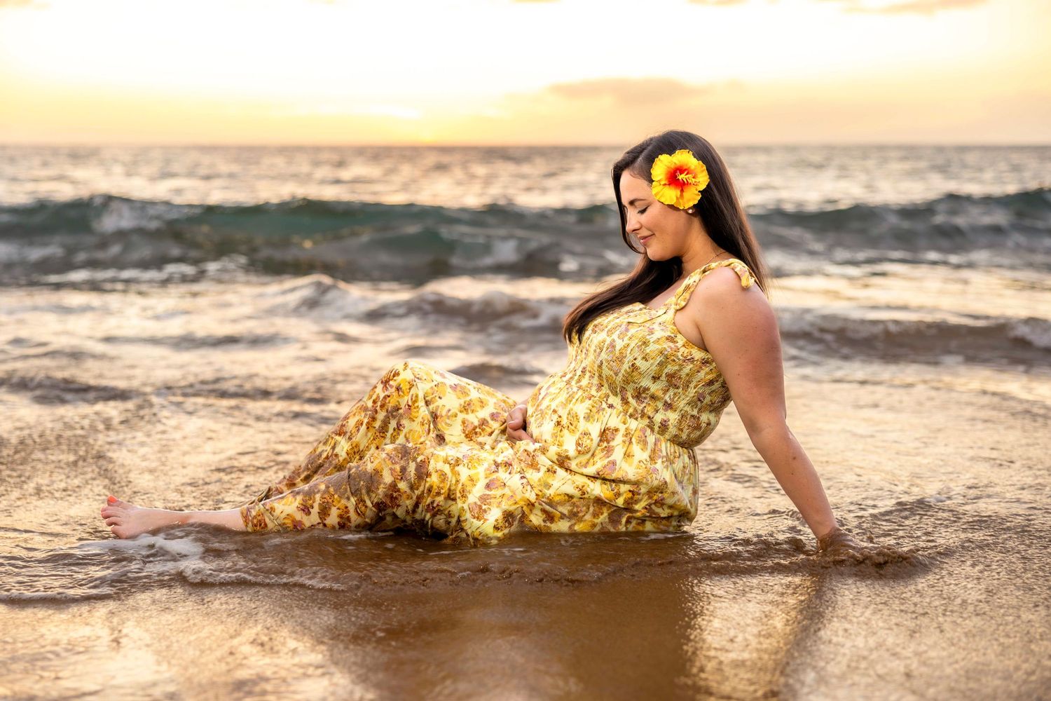 Woman in a yellow floral dress with an orange flower in her hair sits in shallow ocean waves on the beach at golden sunset. Wailea