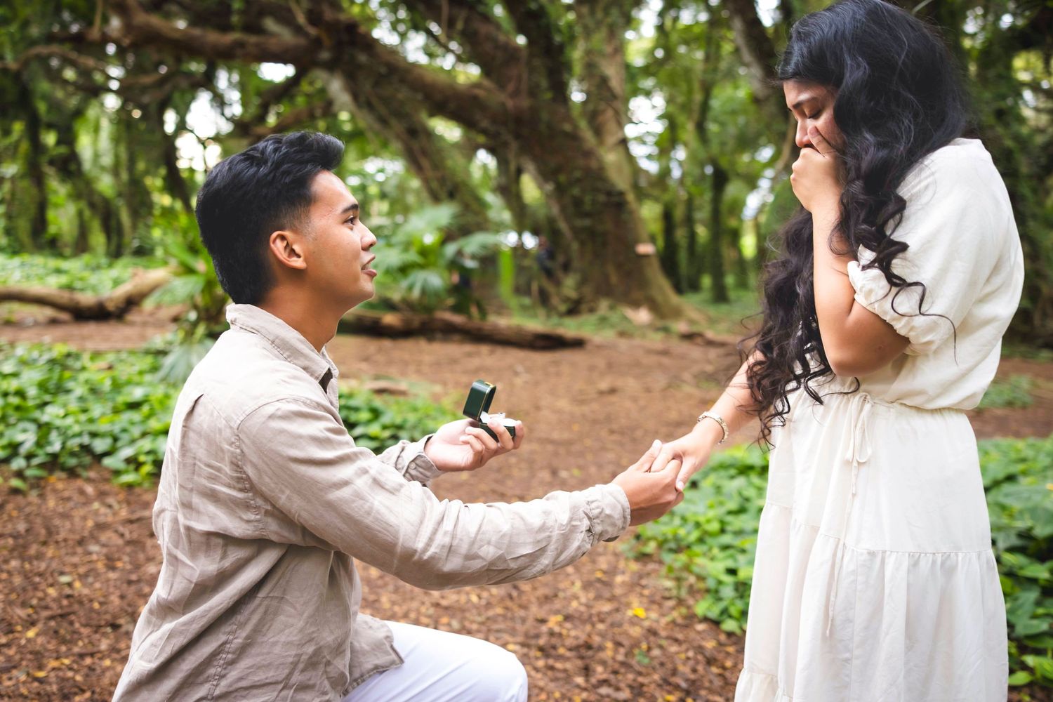A man in a beige shirt kneels on one knee holding a ring box as a surprised woman in a white dress covers her mouth in a leafy park. Honolua