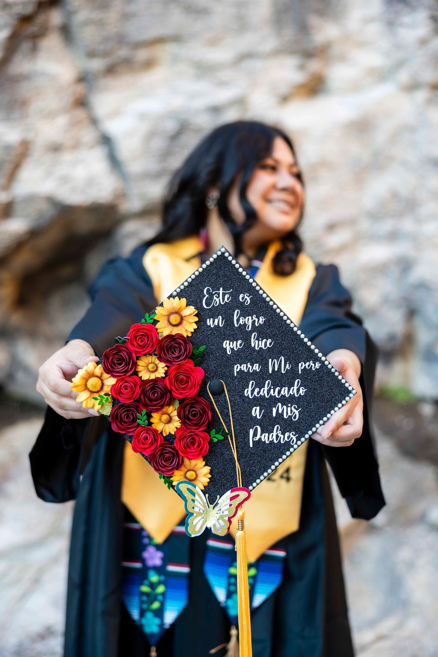 Smiling graduate in black gown and gold stole holds a decorated cap with red paper flowers and a Spanish dedication to her parents.