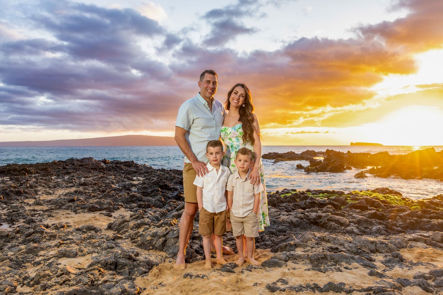 Family of four poses on black lava rocks at sunset with a vibrant purple and orange sky over the ocean in Maui.