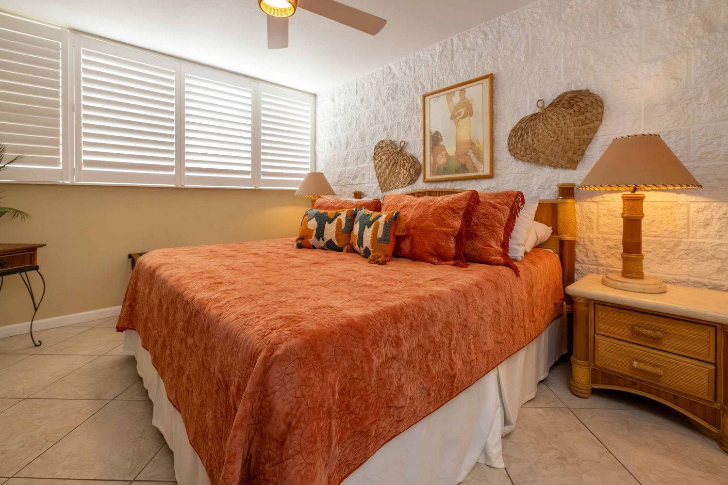 Cozy bedroom with a coral bedspread, decorative pillows, wooden nightstand with lamp, white plantation shutters, and tile flooring.