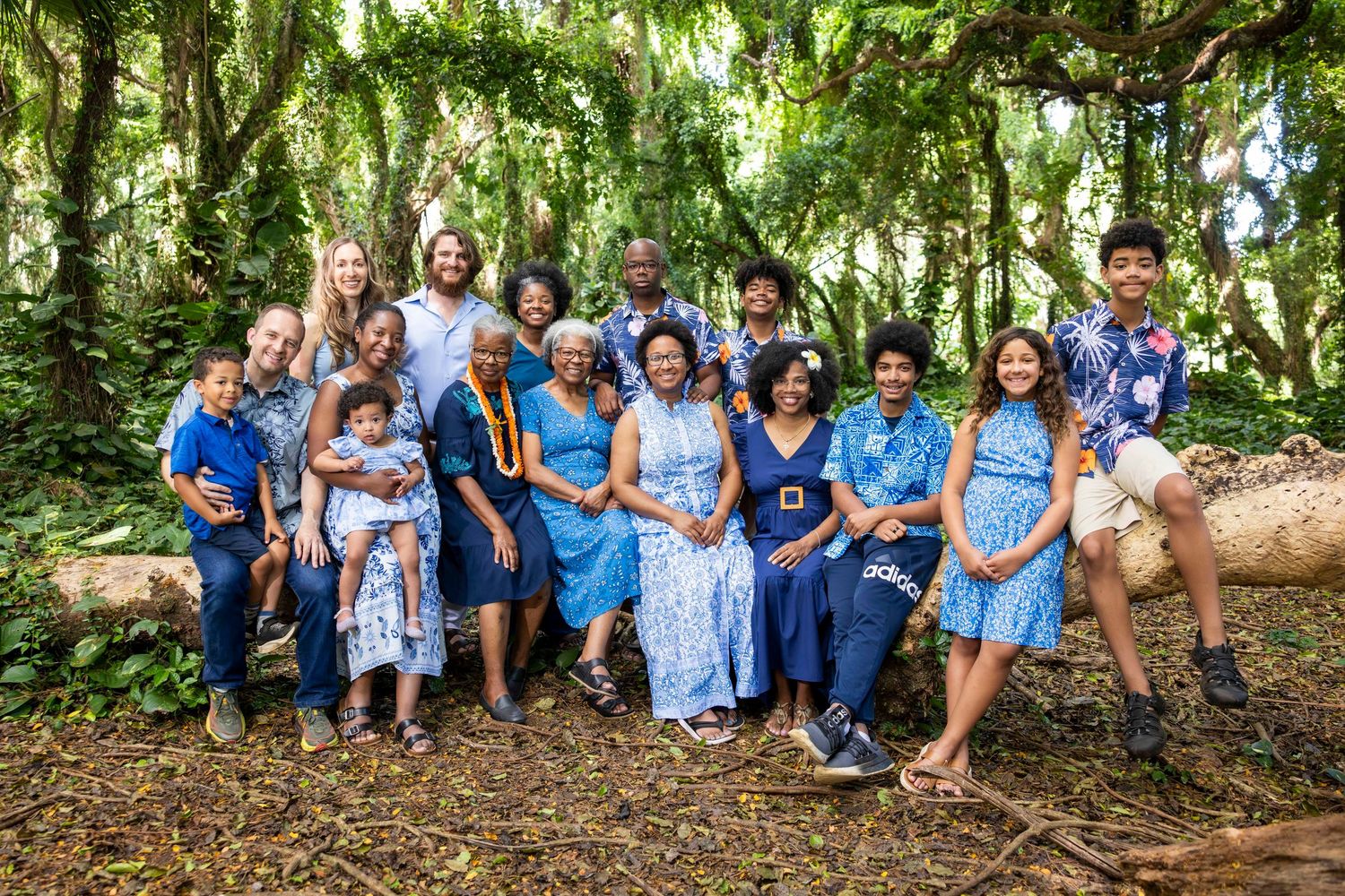 Large multigenerational family in coordinated blue outfits posing together on a log in a lush green tropical forest.