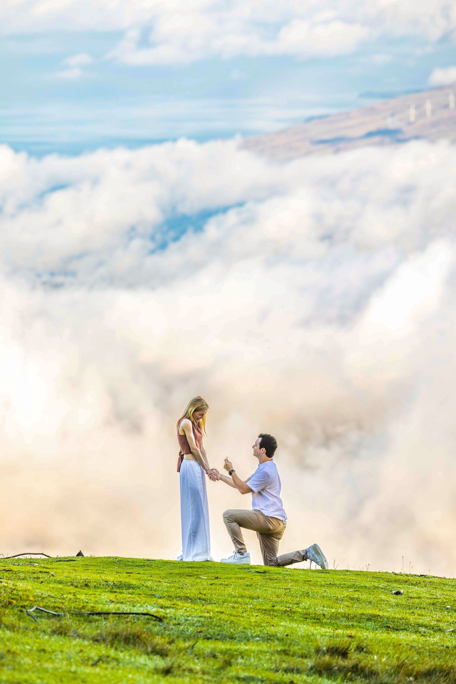 Man kneeling on one knee proposes to a woman in a long dress on a green hilltop above a sea of fluffy white clouds. Poli Poli