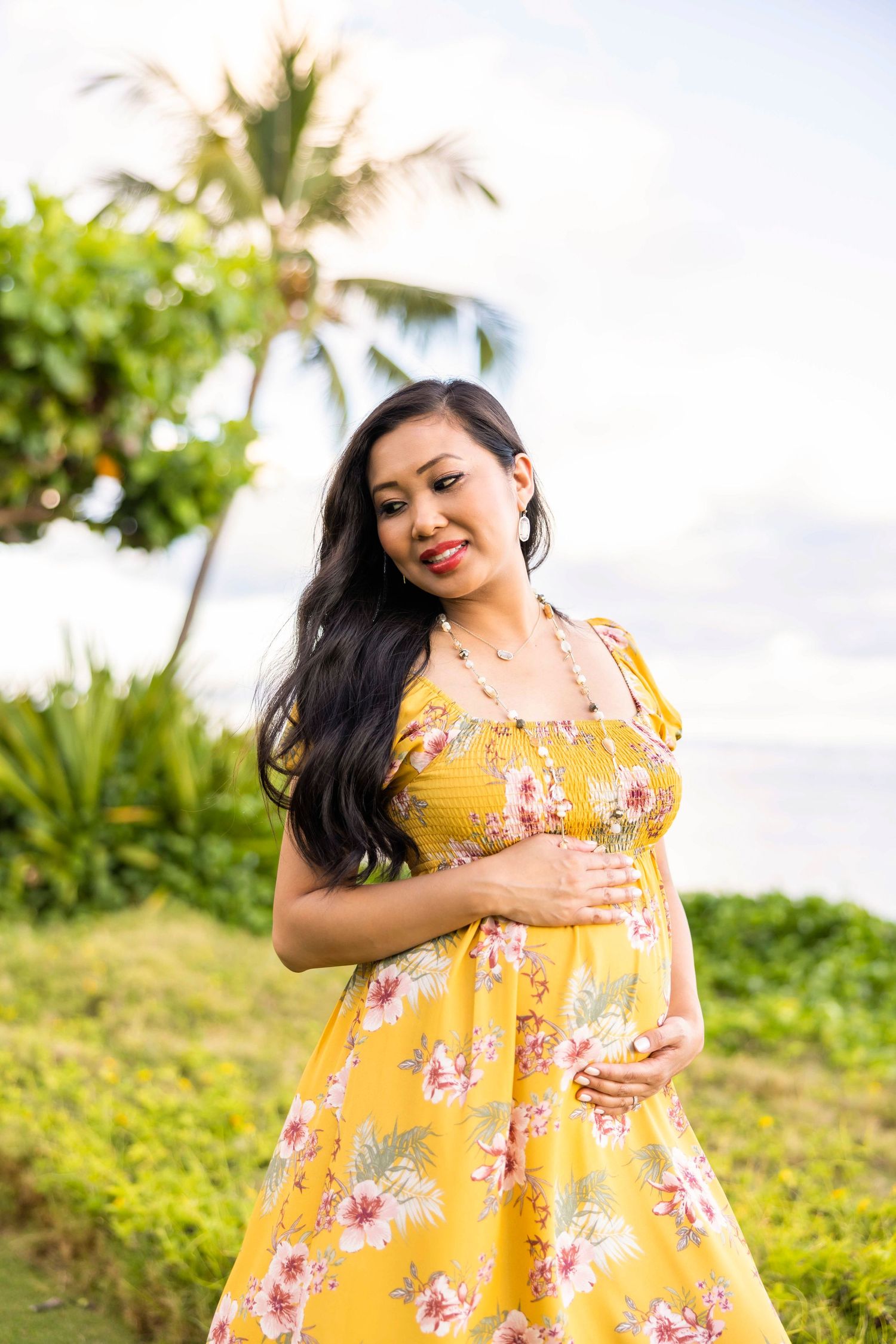 Pregnant woman in a yellow floral dress cradles her baby bump while standing on a grassy hillside with palm trees and ocean behind her.