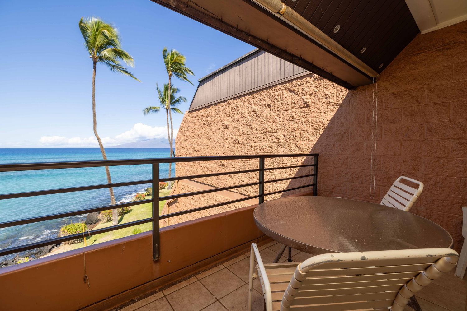 Oceanfront condo balcony with a round table and chairs, palm trees, and sweeping blue Pacific views under a clear sky.