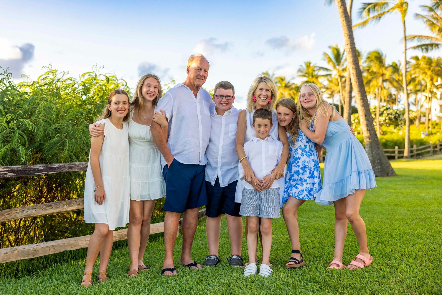Large family of nine in coordinated casual outfits poses on a lush green lawn with palm trees in a tropical Hawaii setting.