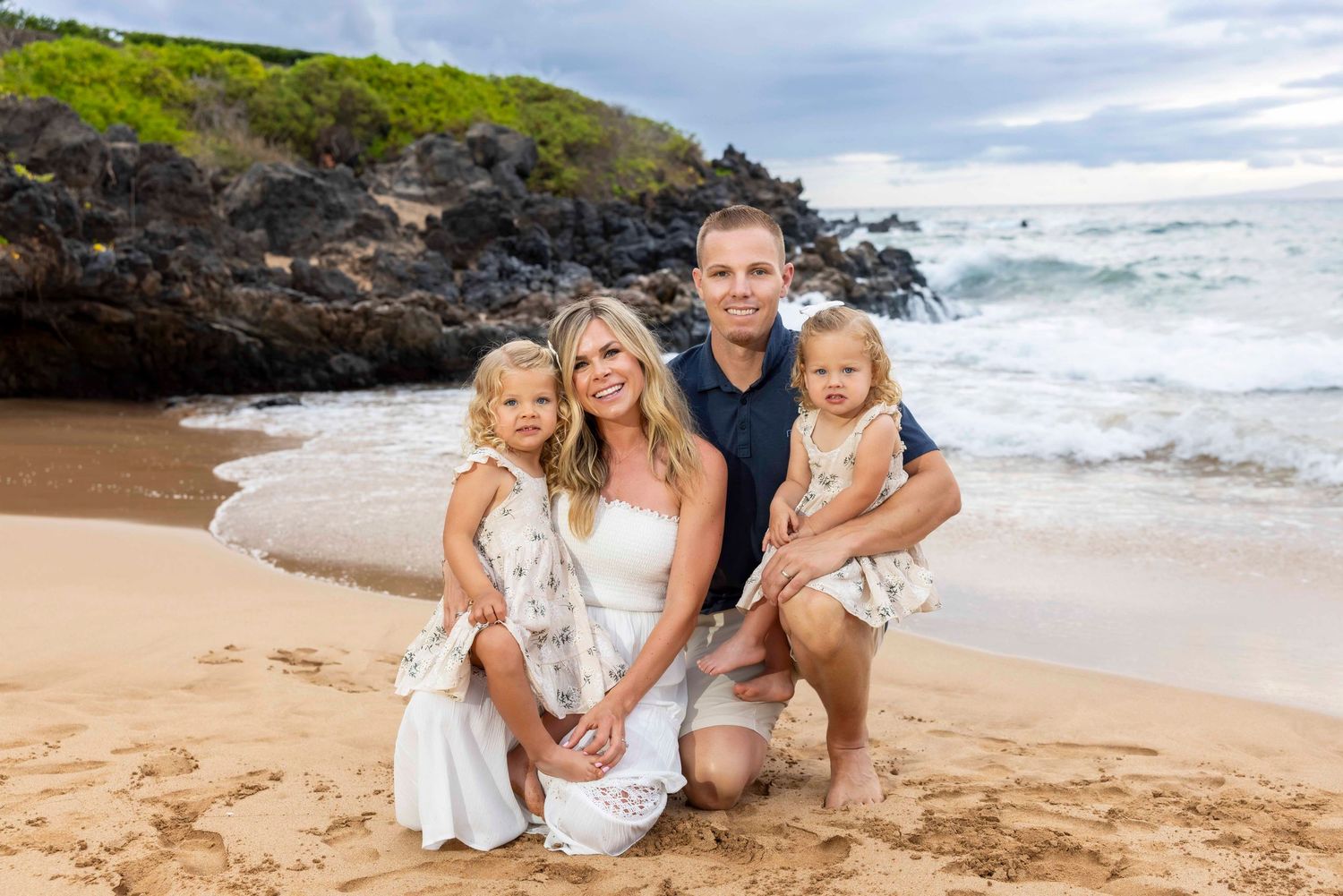 Smiling family of four in white and navy outfits posing together on a sandy beach with ocean waves and dark lava rocks behind them.