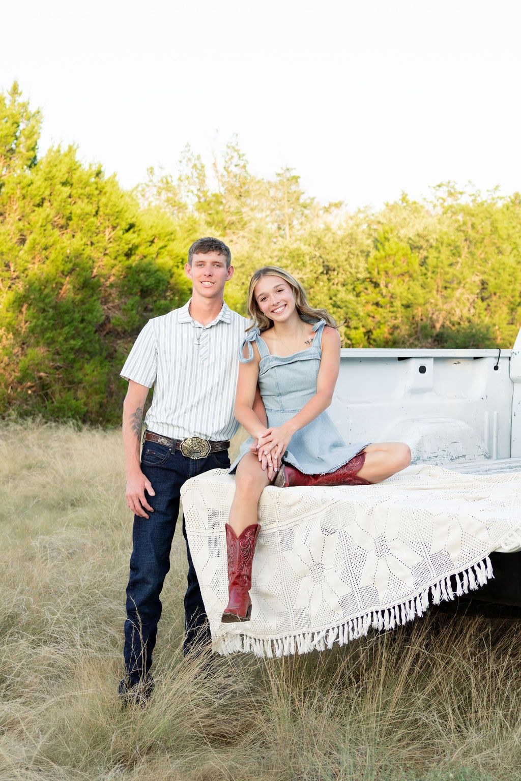 A young couple poses by a white truck bed in a field, she wears boots and a blue dress, he wears jeans and a striped shirt.