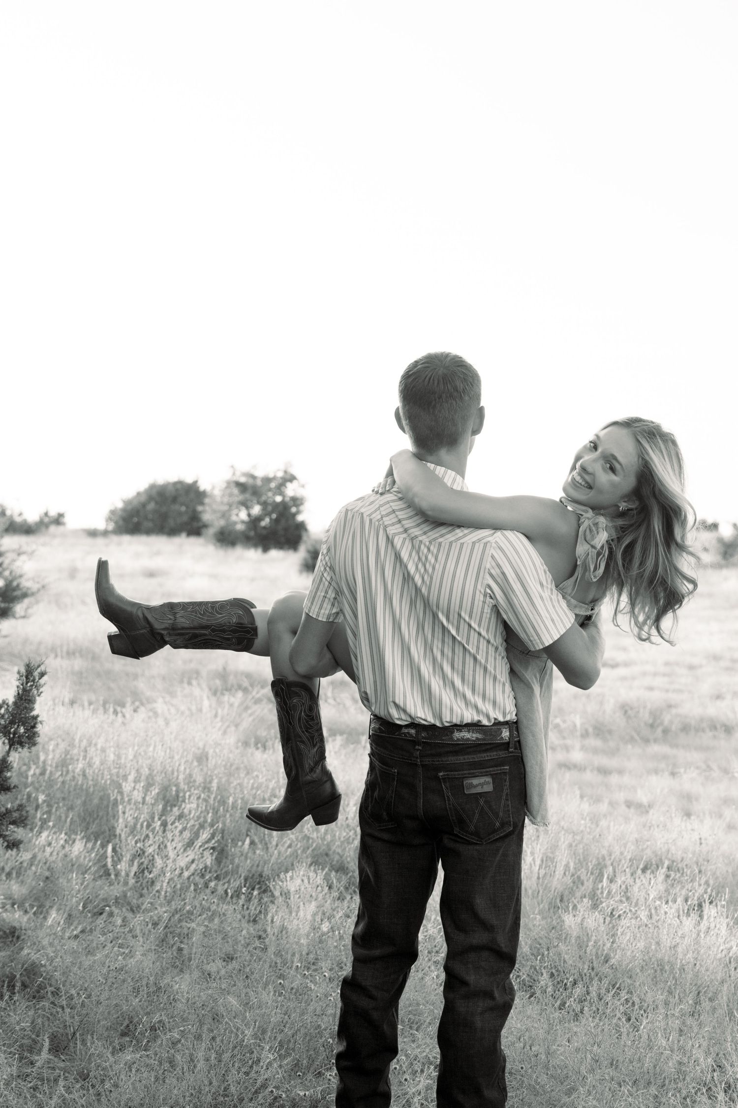 Black and white photo of a man carrying a woman in cowboy boots through a rural field.