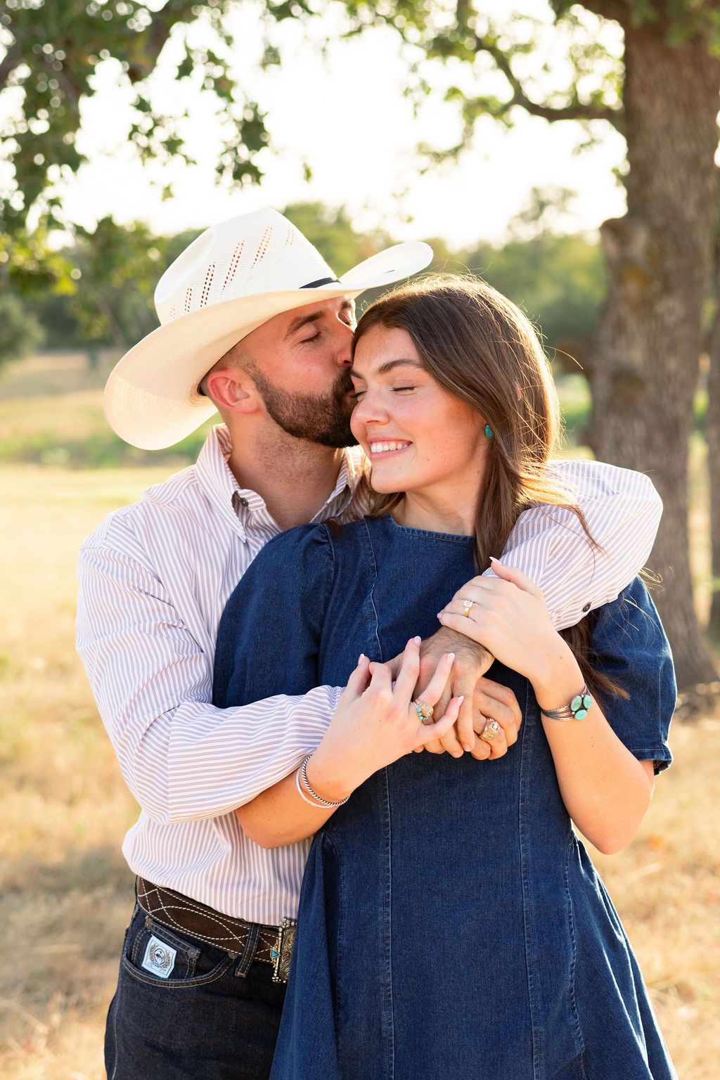 A couple embraces outdoors, man in cowboy hat kissing woman's cheek as she smiles, golden hour light filtering through trees.