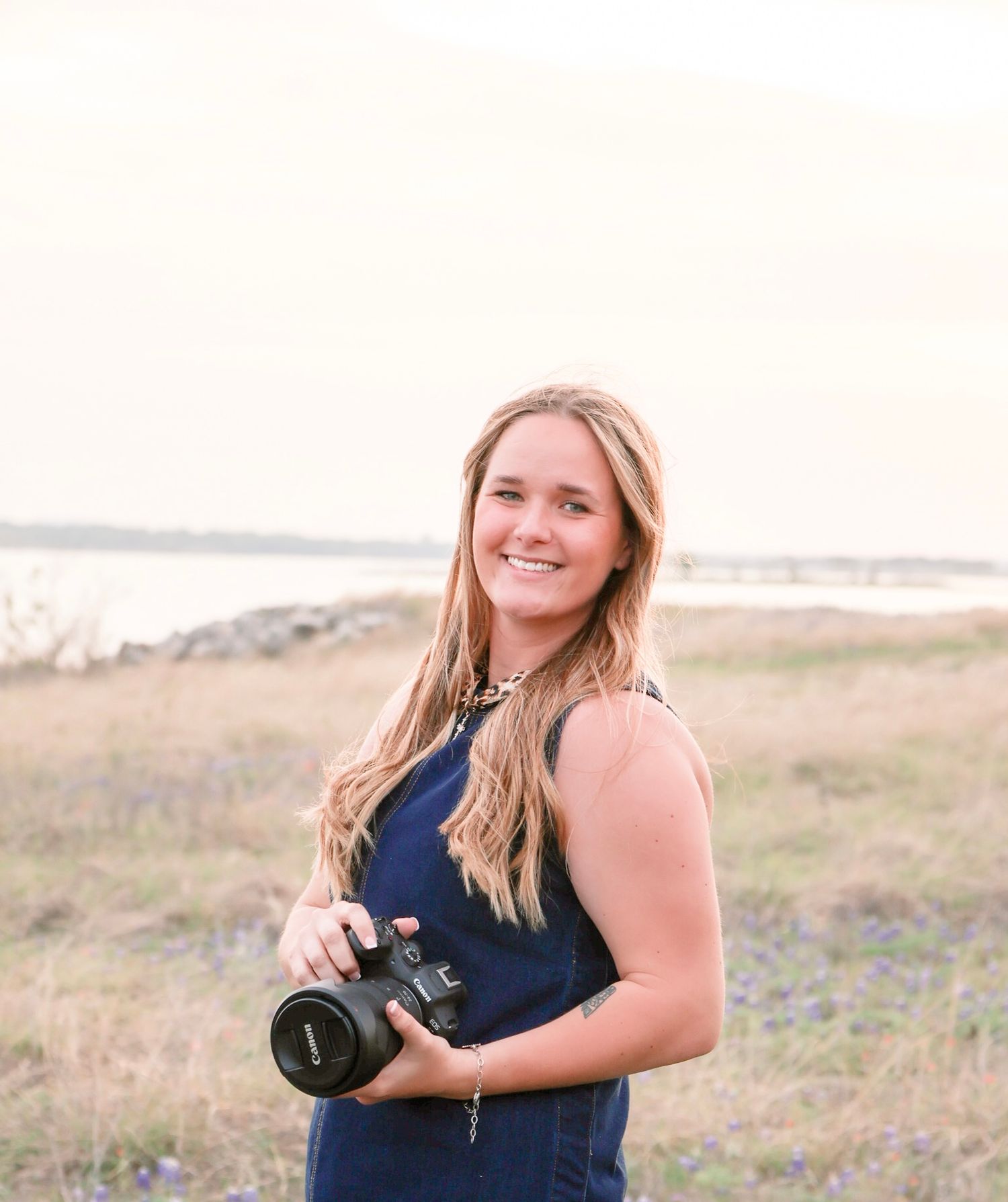 Smiling young woman holding DSLR camera standing in golden field near waterfront at sunset.