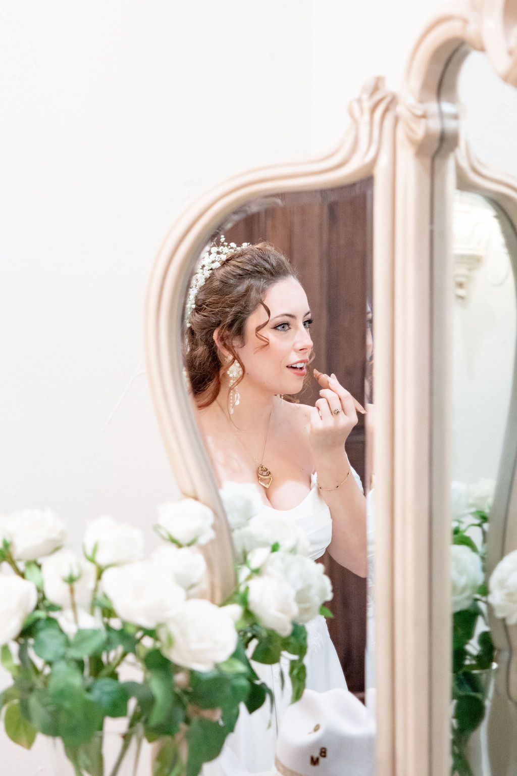 Bride applying lipstick reflected in ornate mirror surrounded by white roses while getting ready.