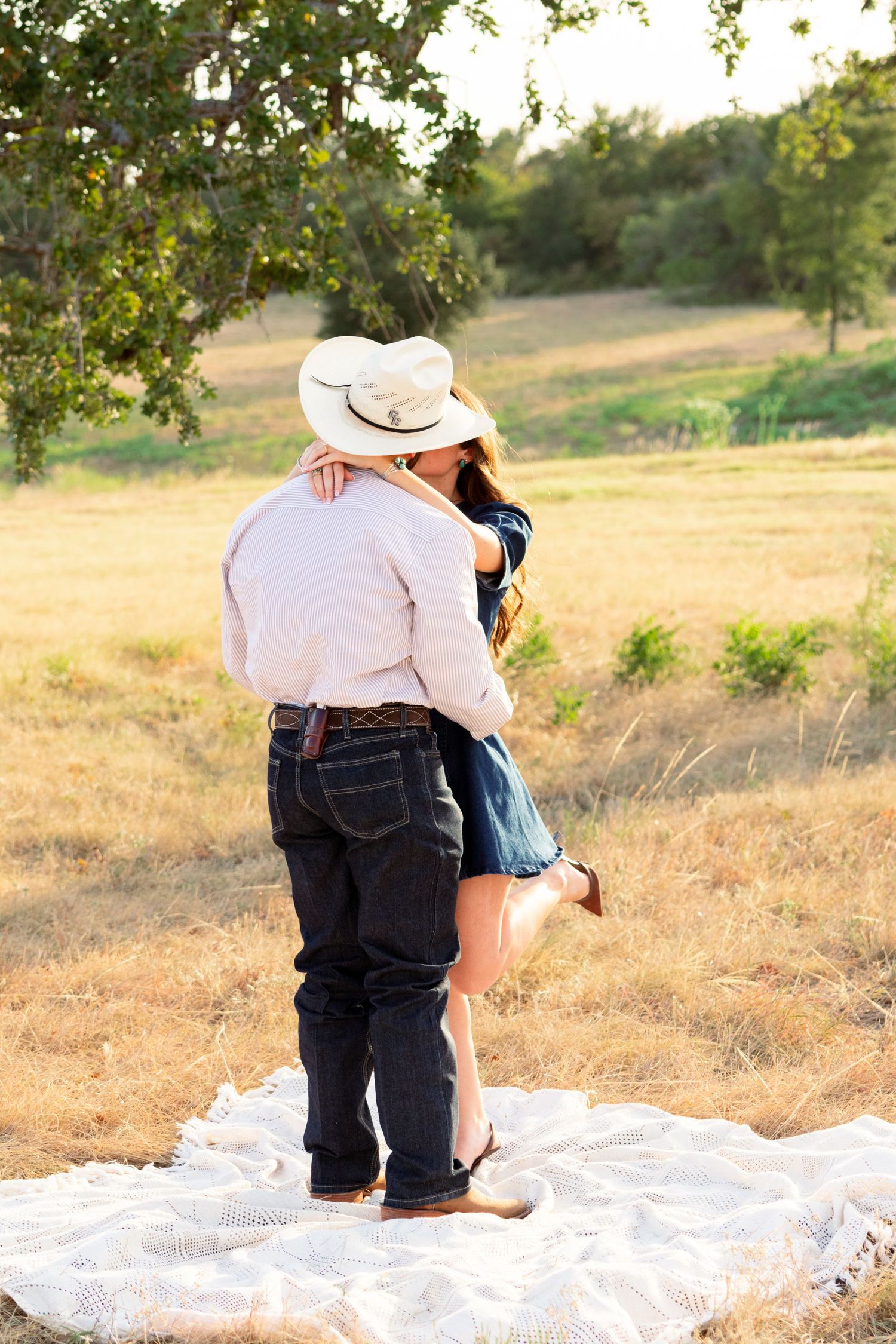 Couple embracing and kissing on a blanket in a sunlit field, man in cowboy hat and woman in blue dress lifting her leg.