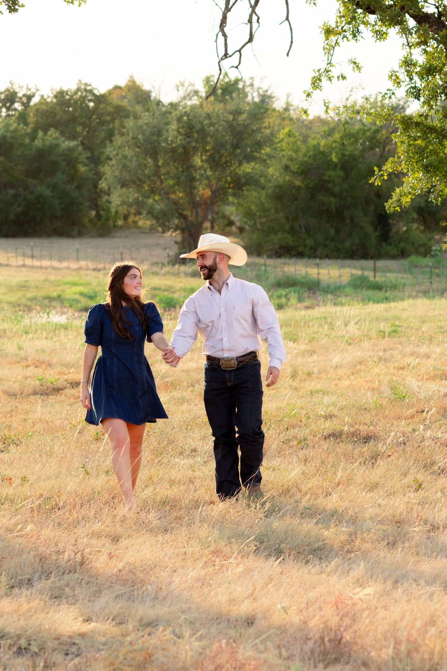 A couple in western attire holds hands walking through a golden field, man in cowboy hat and woman in blue dress.