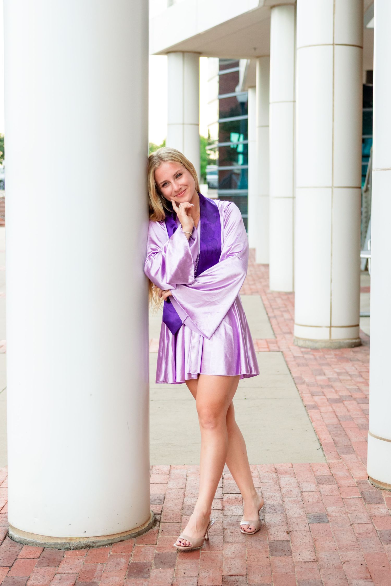 Smiling blonde woman in purple graduation gown and heels posing beside white columns on a brick walkway.