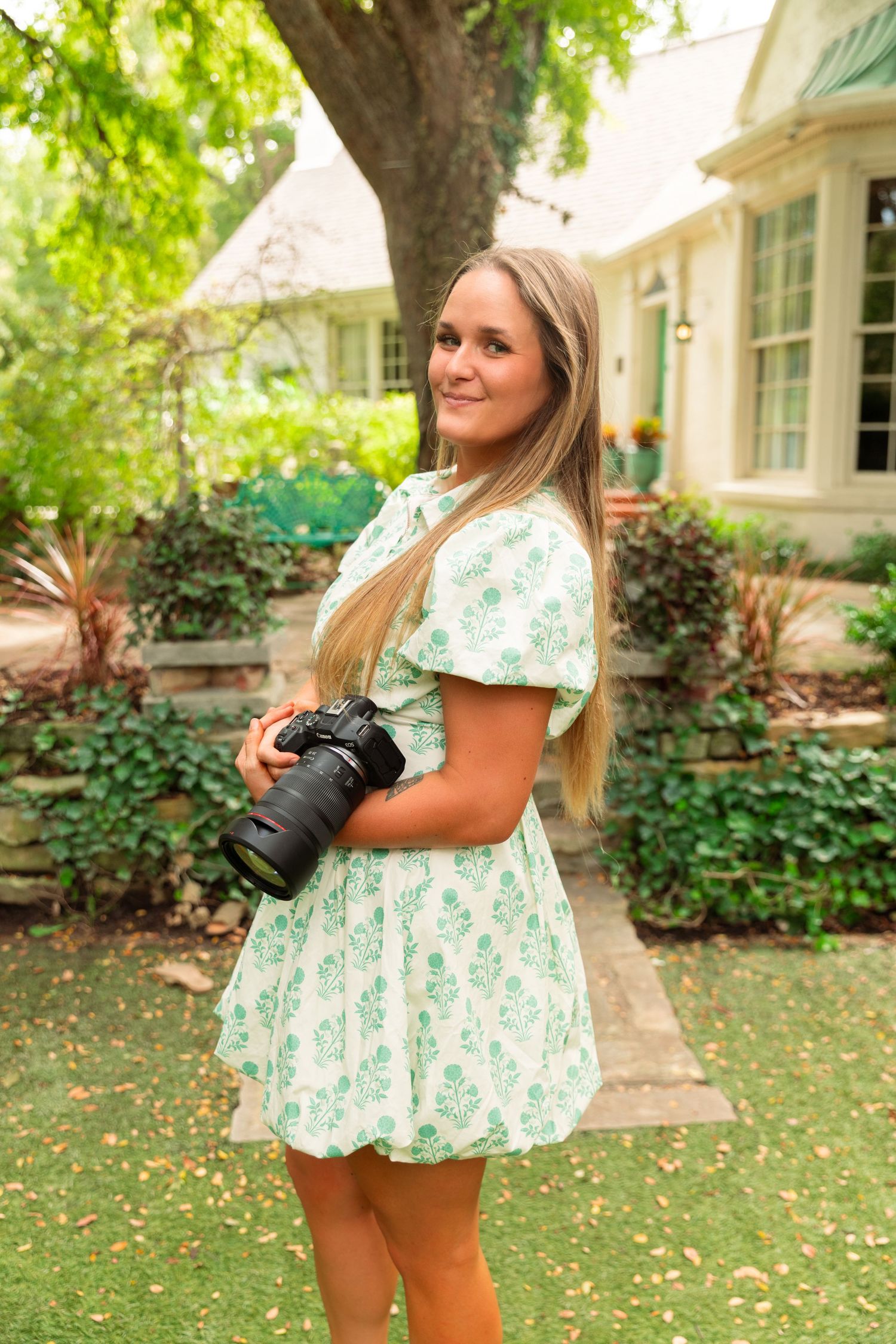 Young blonde woman in floral green dress holding DSLR camera, posing outdoors near lush garden and house.