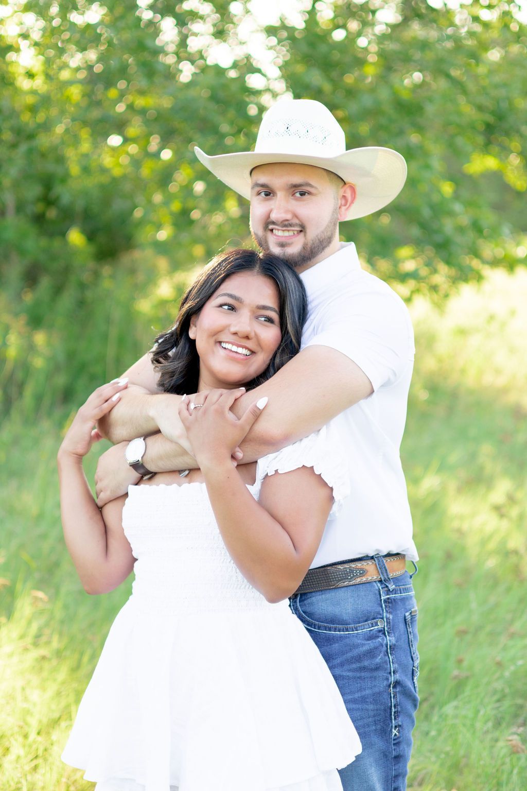 A smiling couple poses outdoors, man in cowboy hat and white shirt hugging woman in white dress amid lush greenery.