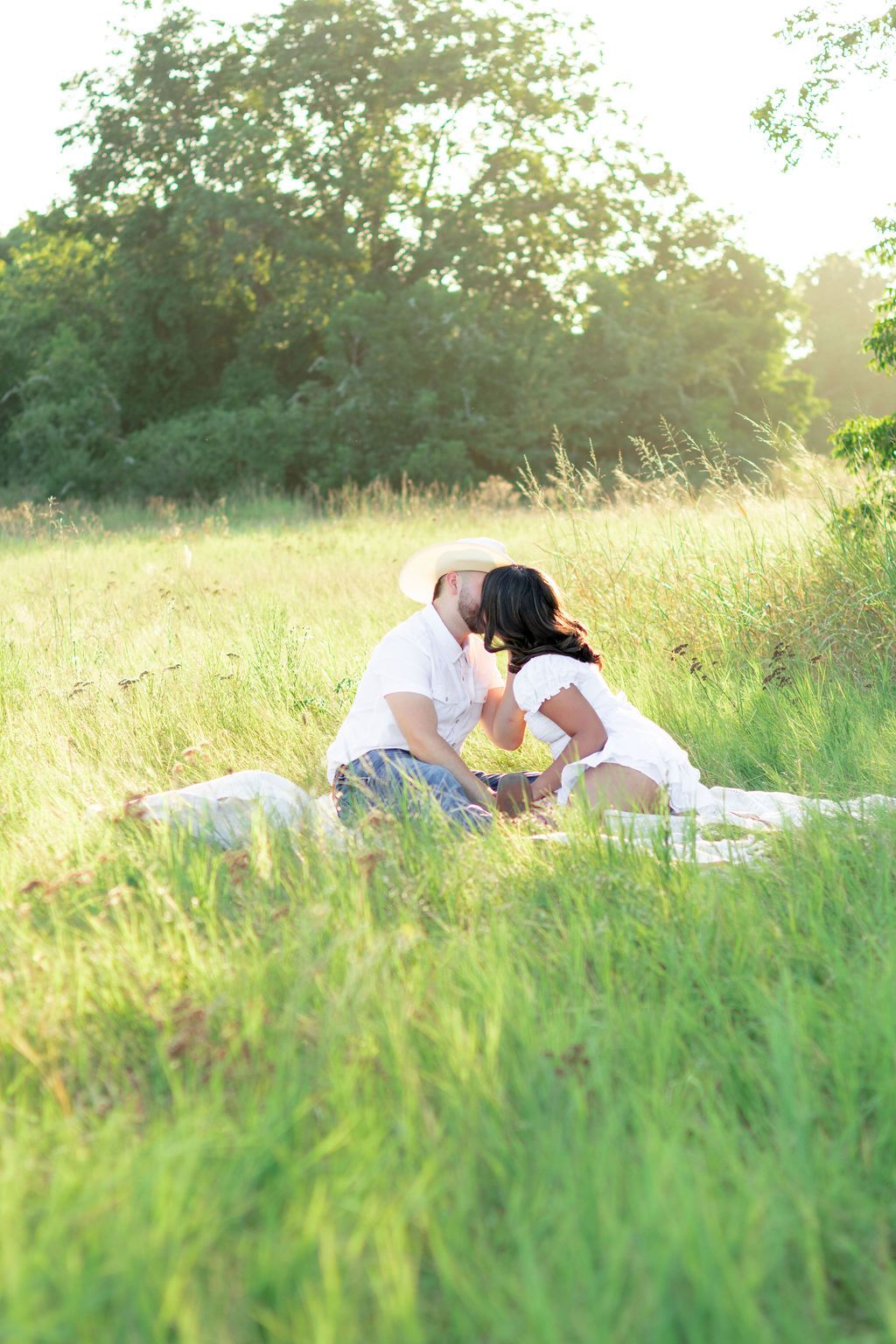 Couple sharing a romantic kiss while sitting on a blanket in a sunny green meadow surrounded by tall grass and trees.