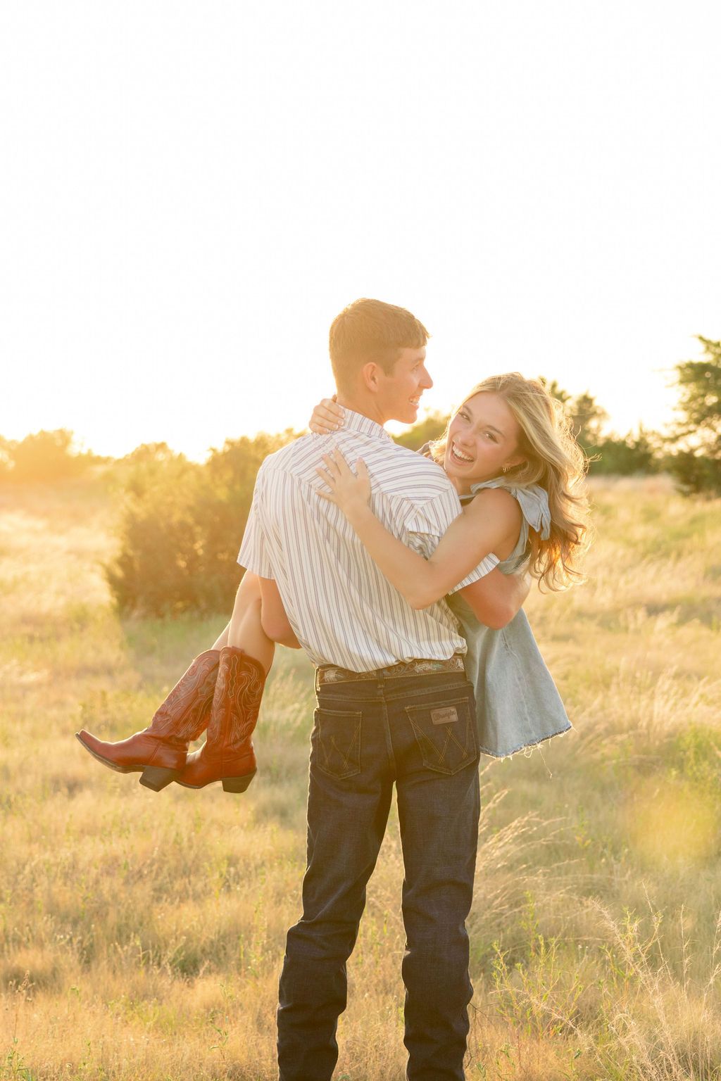 Couple embracing at golden hour in a field, woman in cowboy boots and denim dress laughing joyfully.