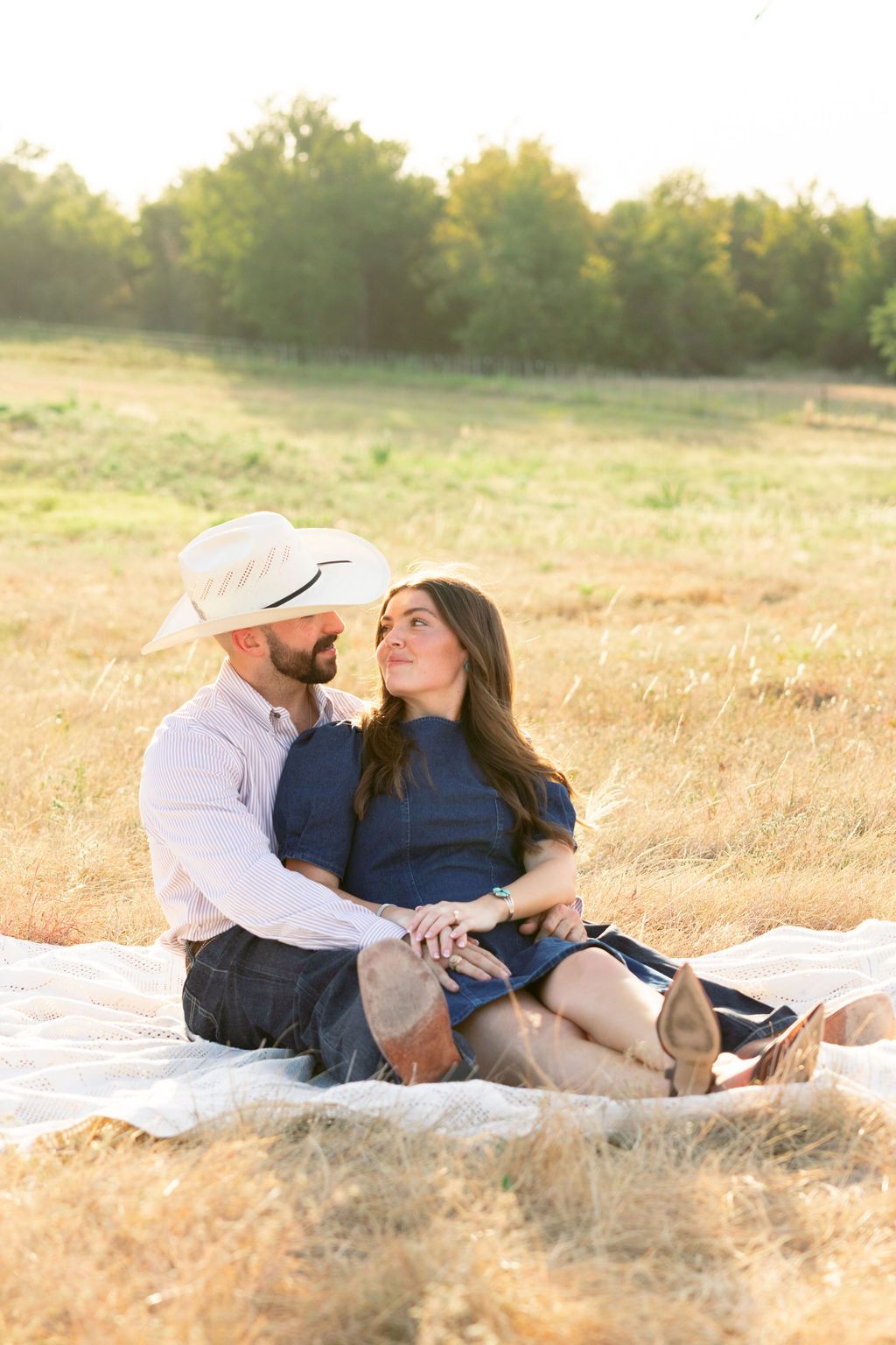 Couple sitting on blanket in golden field, man in cowboy hat embracing woman in blue dress at sunset.