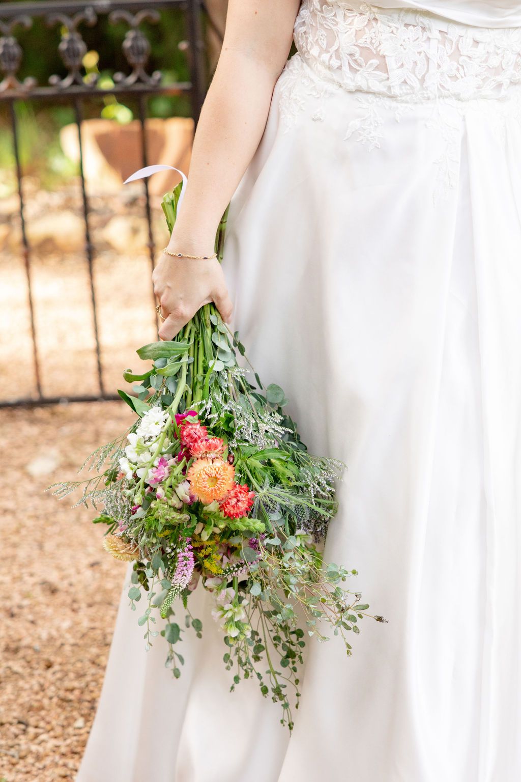 Bride in white lace gown holding a cascading wildflower bouquet with colorful blooms and greenery near iron gate.