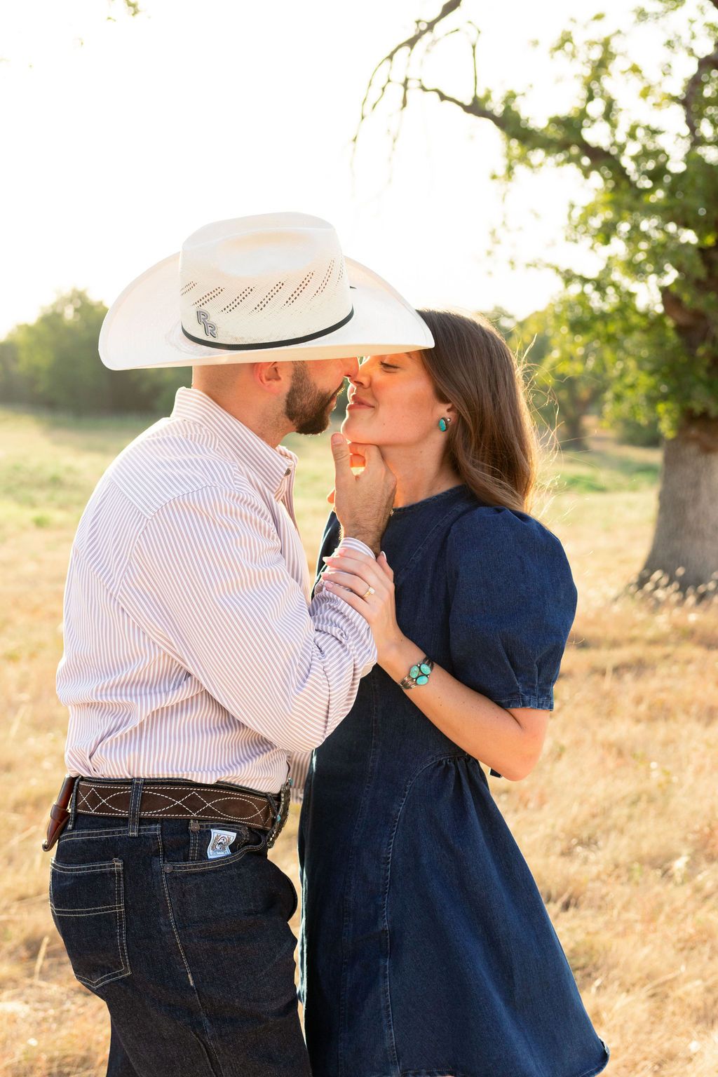 Couple sharing a romantic kiss outdoors, man in white cowboy hat and woman in denim dress, golden hour lighting.