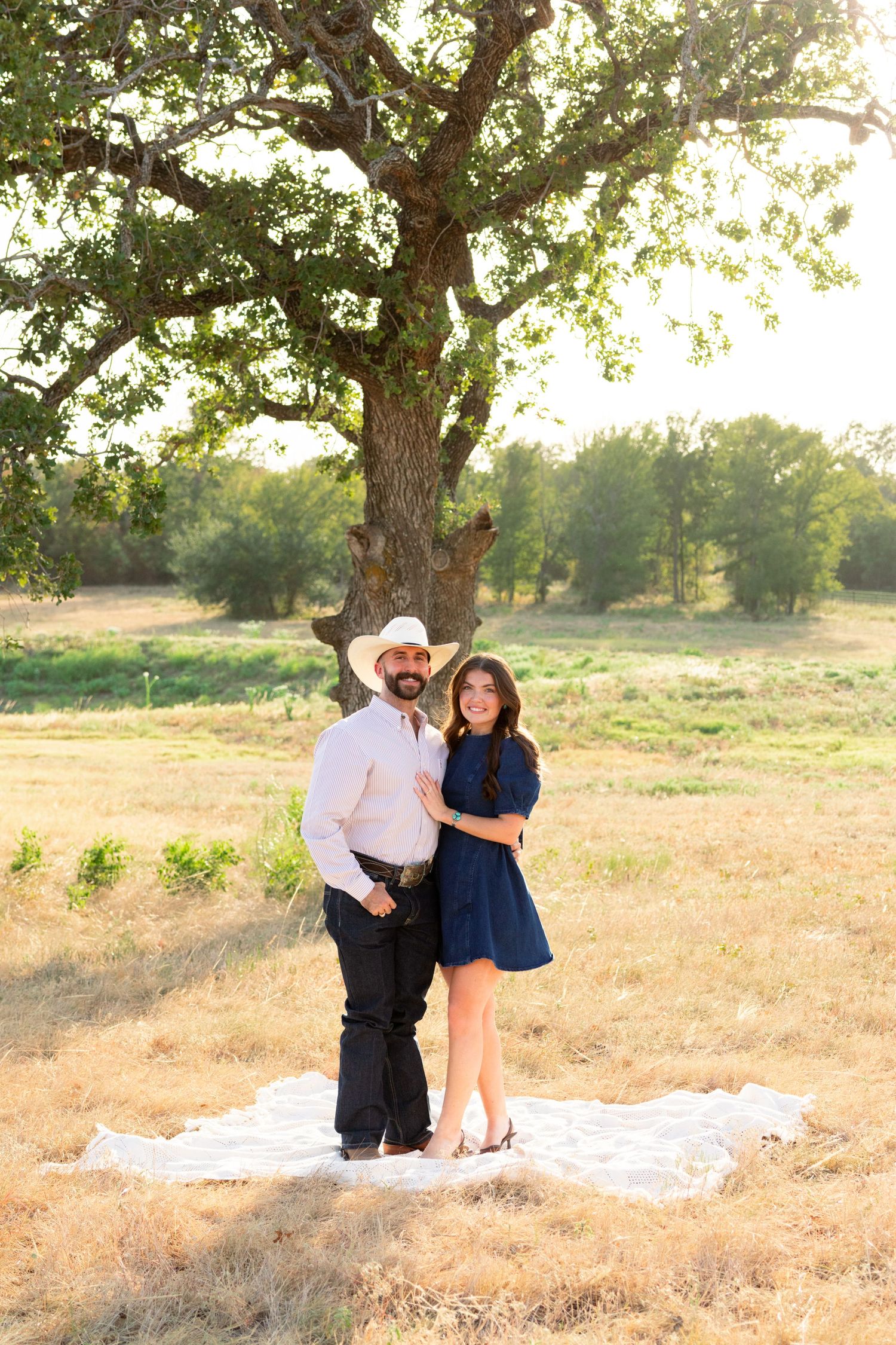 Couple posing together on a blanket in a golden field beneath a large oak tree, man wearing cowboy hat and woman in navy dress.